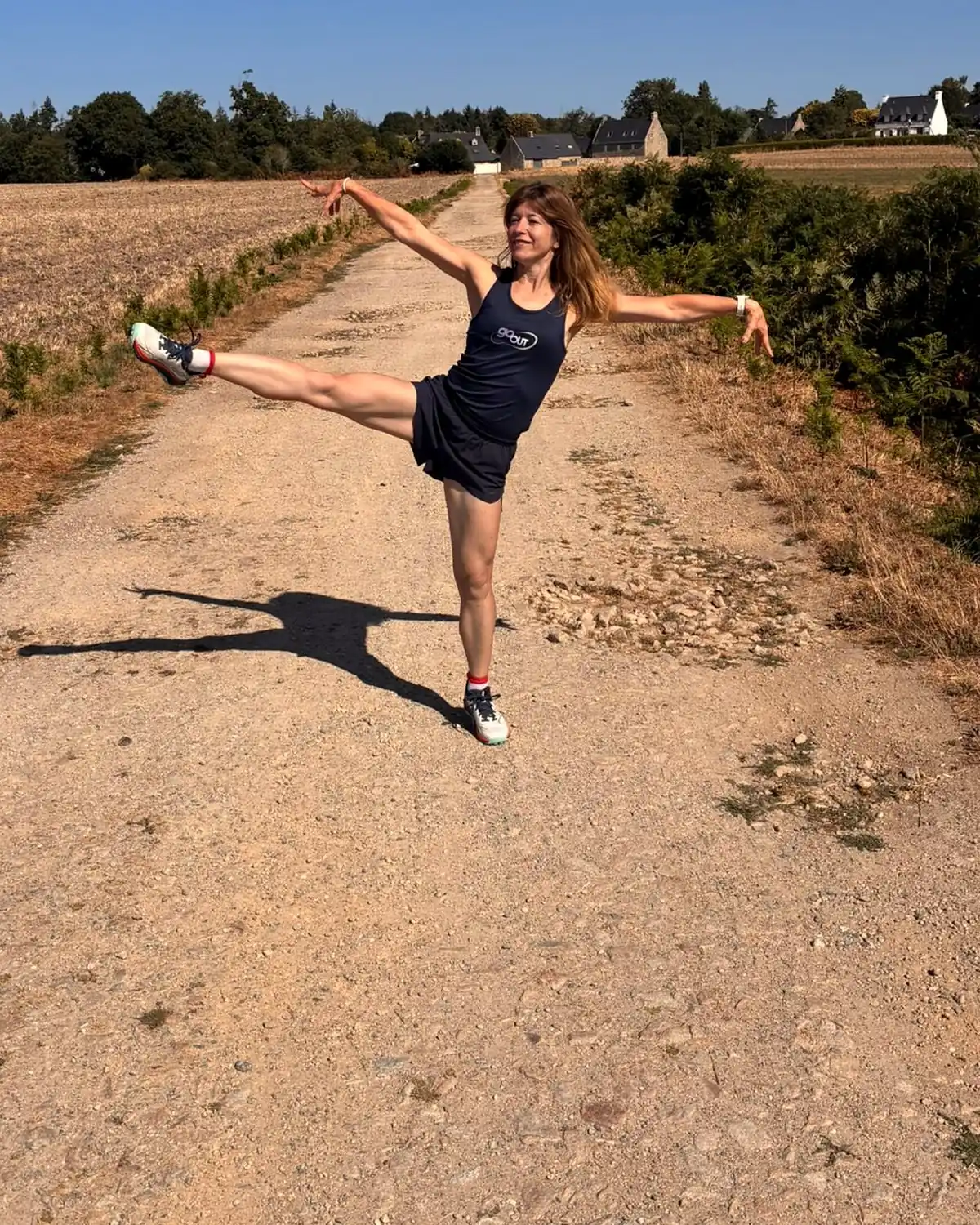 Une femme en tenue Go Out, bras et jambe tendus, sur un chemin de campagne en Bretagne