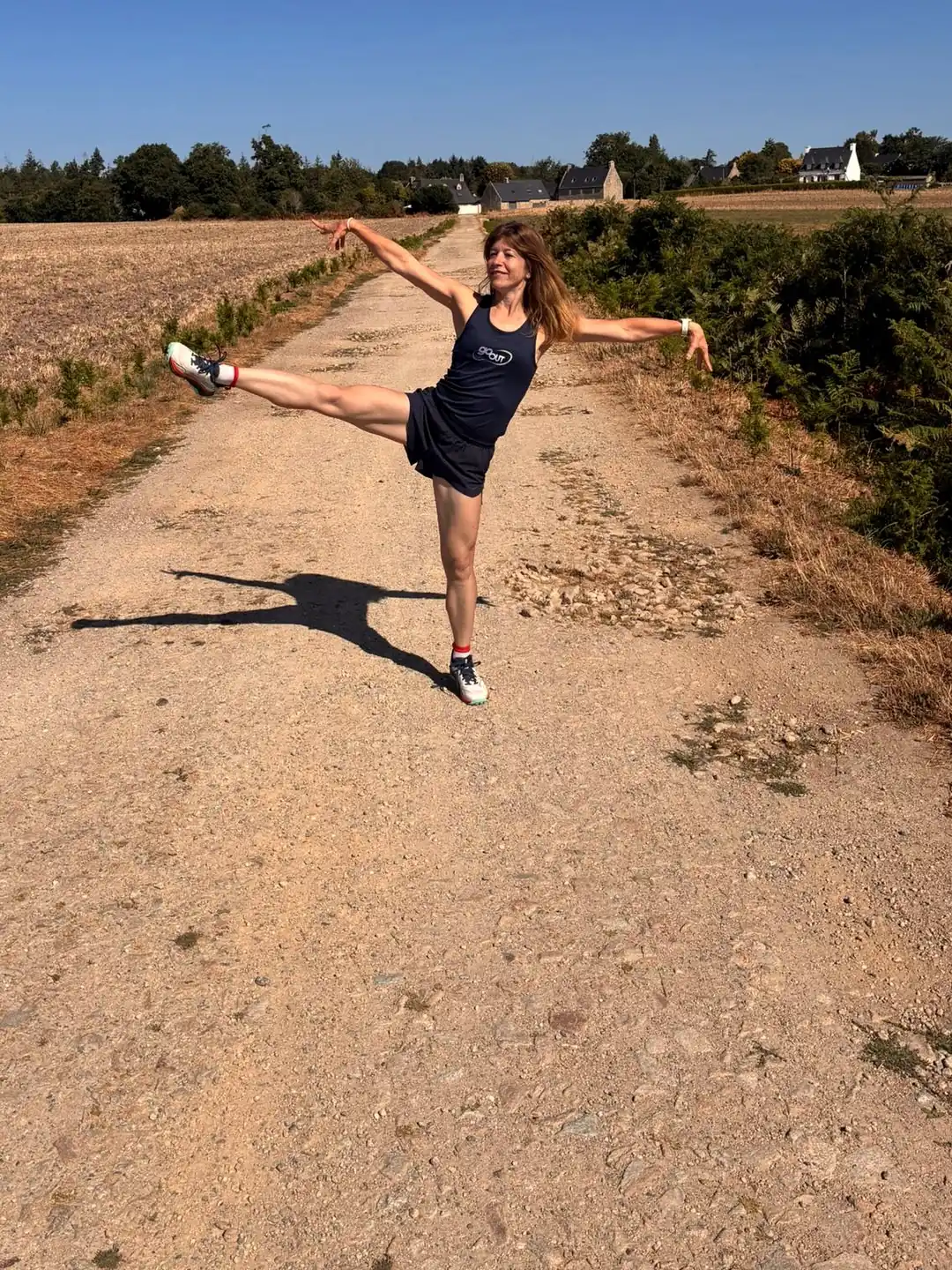 Une femme en tenue Go Out, bras et jambe tendus, sur un chemin de campagne en Bretagne